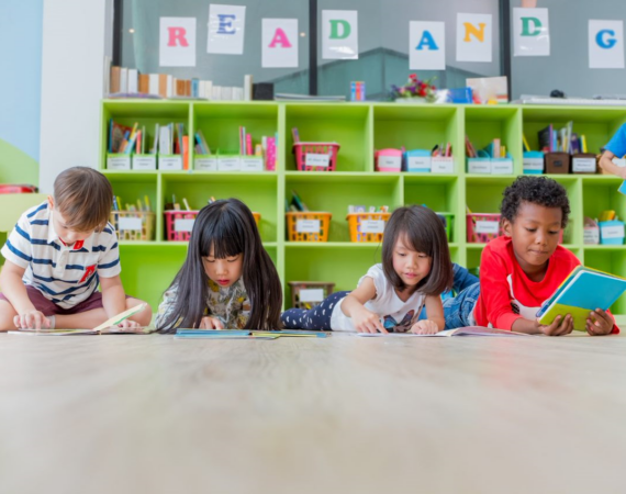 Four children lying on the floor reading books in a classroom