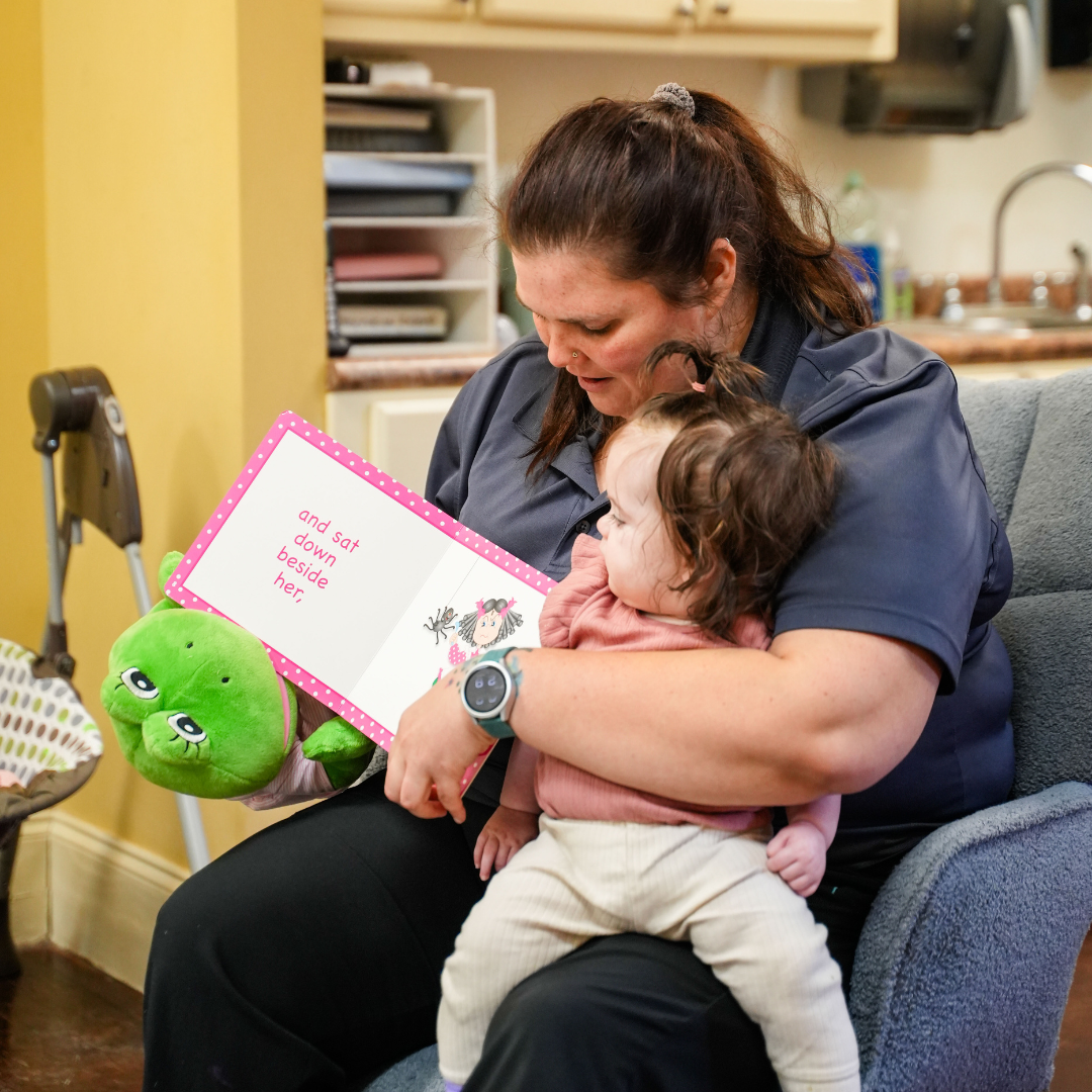 Person holding a child and a book with text "and sat down beside her"