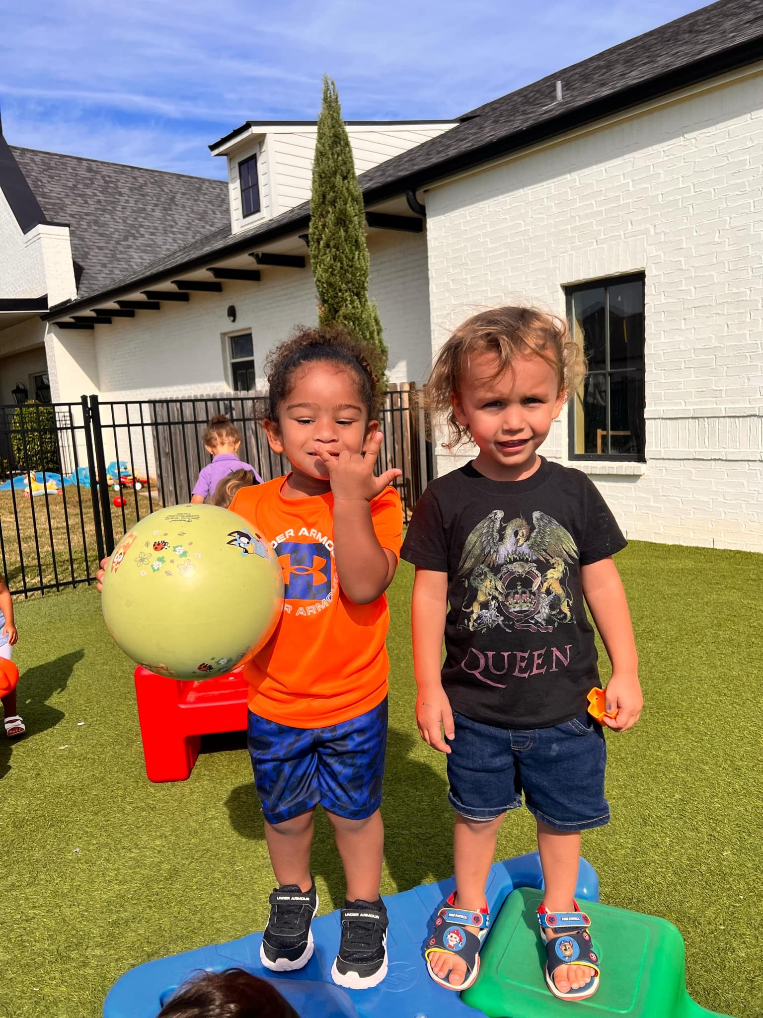 Two children standing on playground equipment, one holding a yellow ball