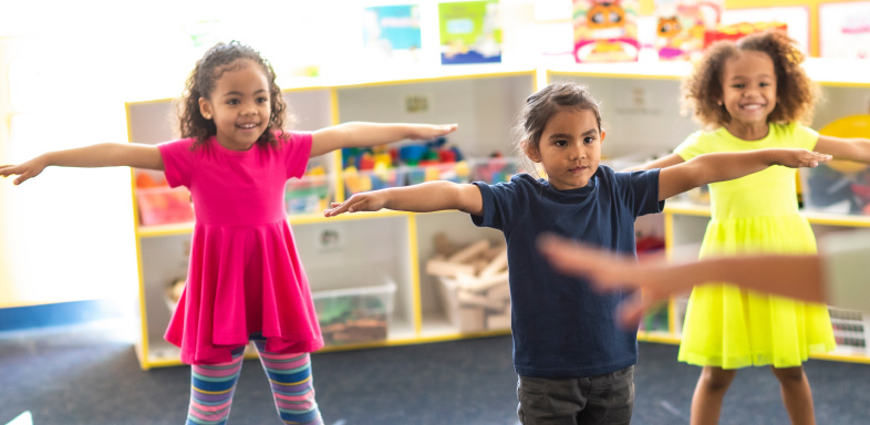 Three children in a classroom standing with arms outstretched
