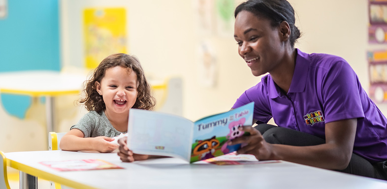 A person reading a book to a child at a table in a classroom
