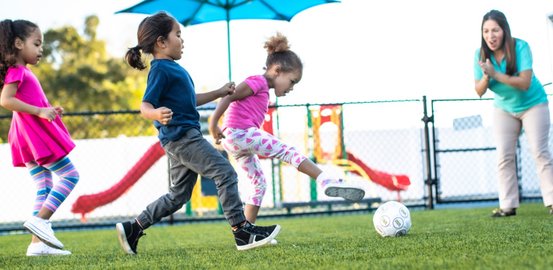 children kicking a soccer ball while an adult watches on a playground