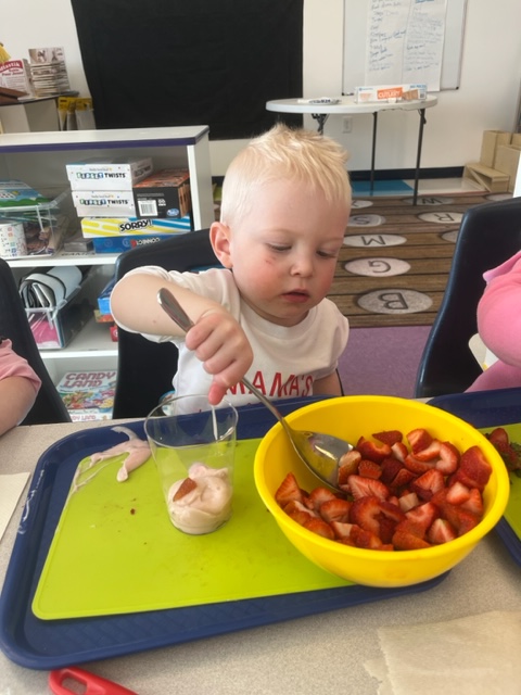 Child using a spoon to mix food in a cup, with a bowl of strawberries nearby