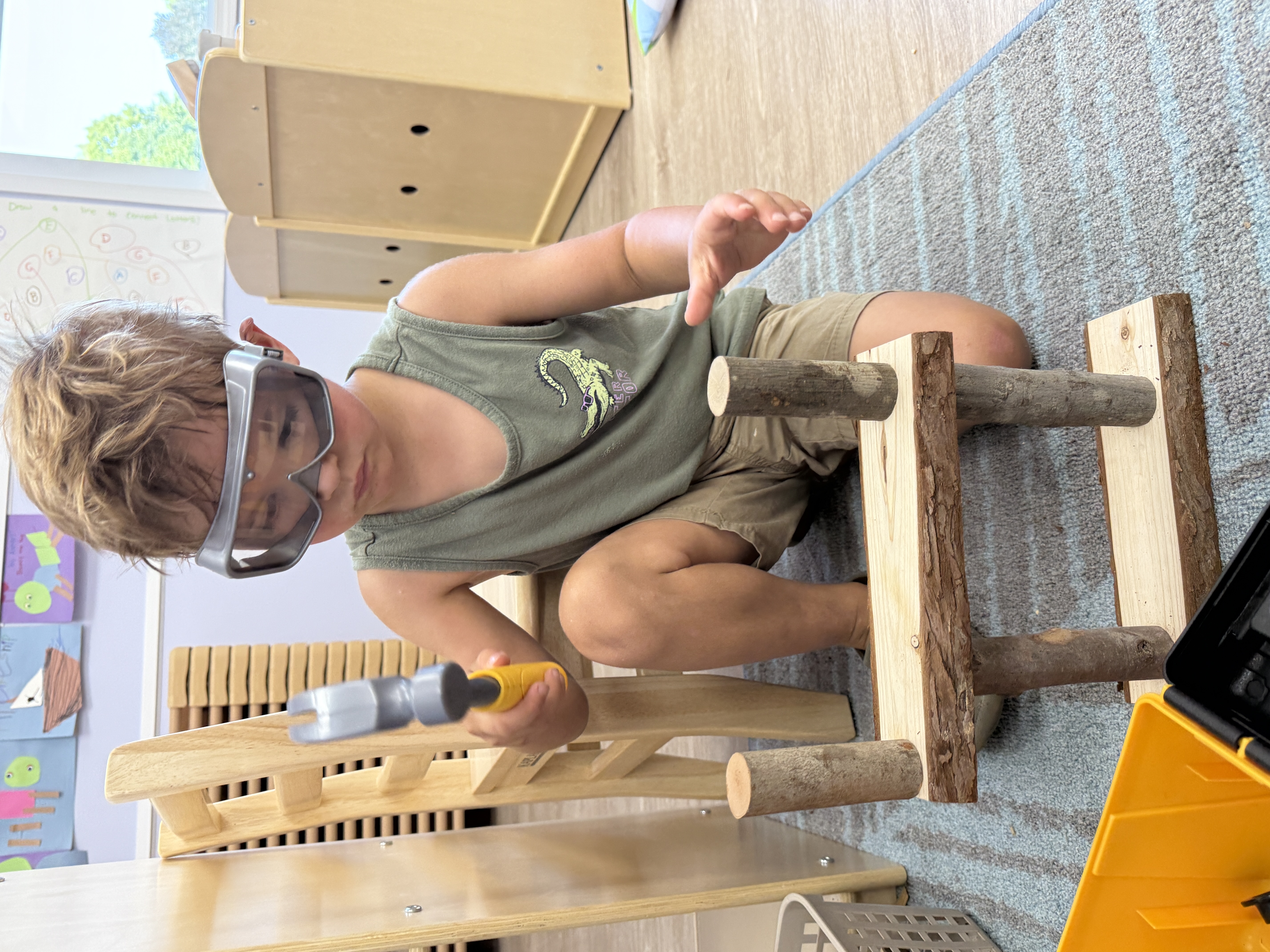 Child wearing safety goggles and holding a toy hammer, building with wood blocks