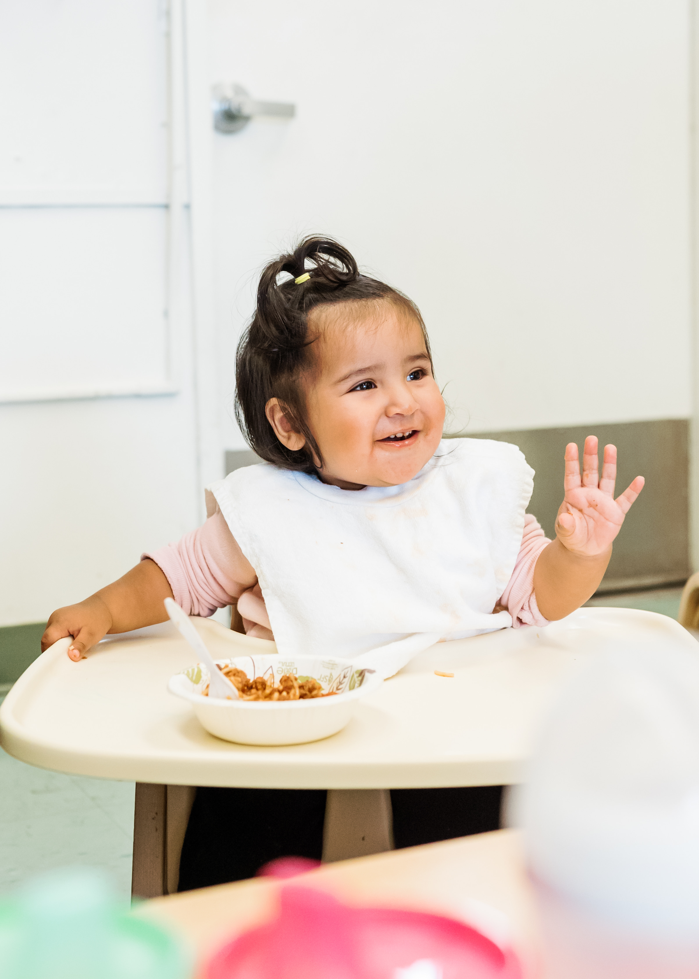 Child in a high chair with a bib, food on the tray, raising a hand