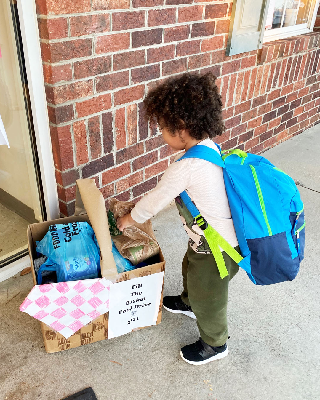 Child with backpack putting items into a box labeled "Fill The Basket Food Drive"