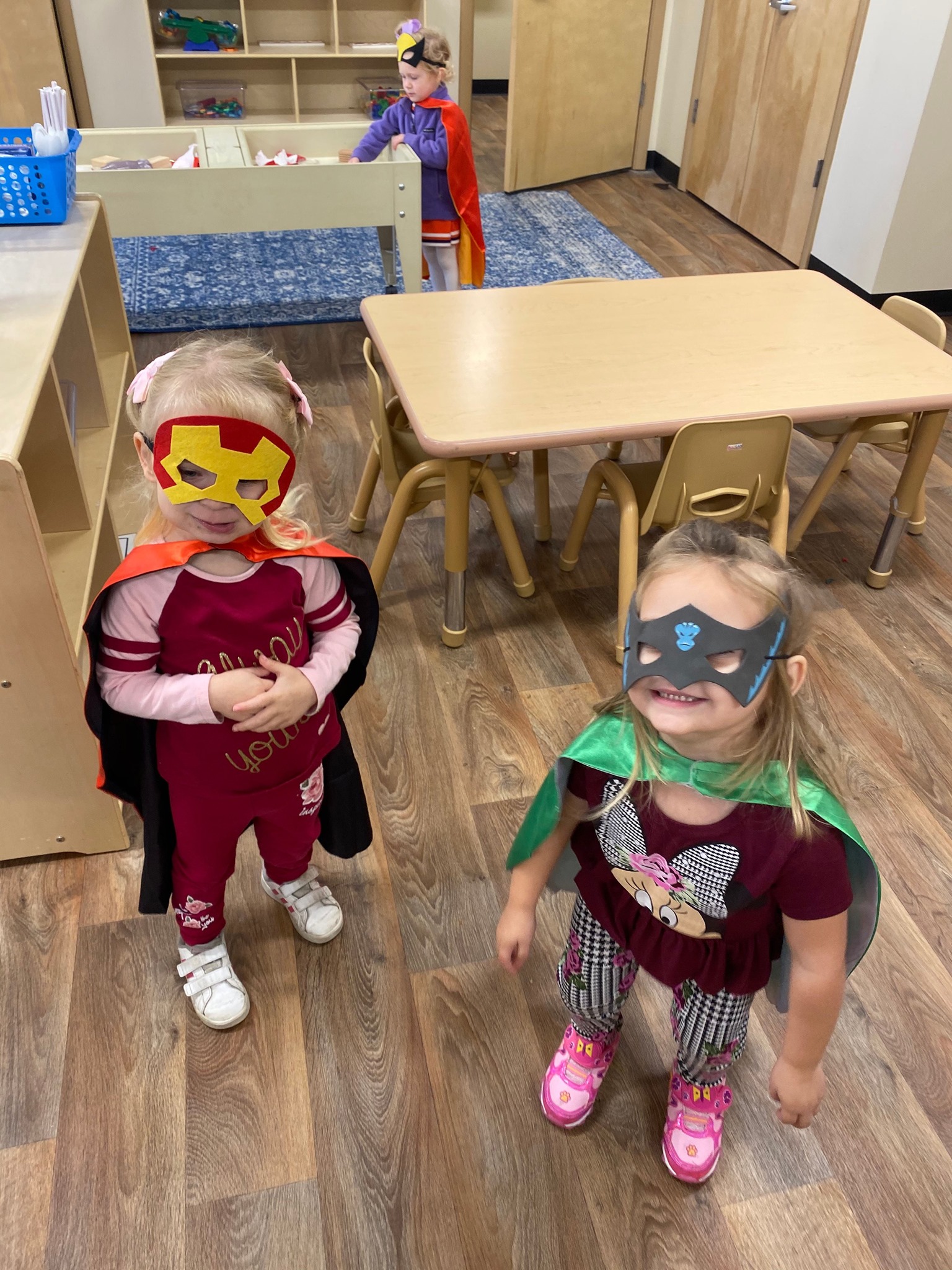 Three children wearing superhero masks and capes in a classroom