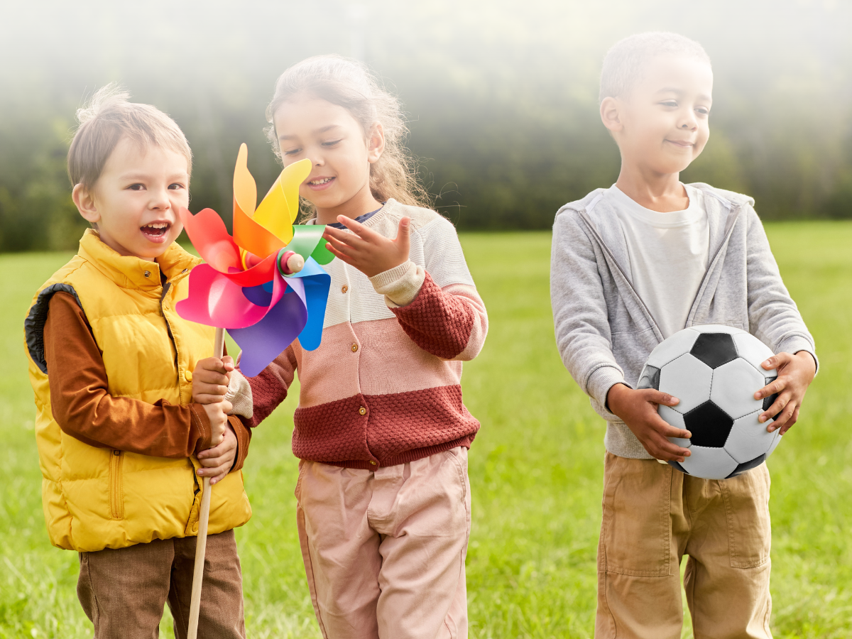 Three children standing in a grassy field with a pinwheel and a soccer ball