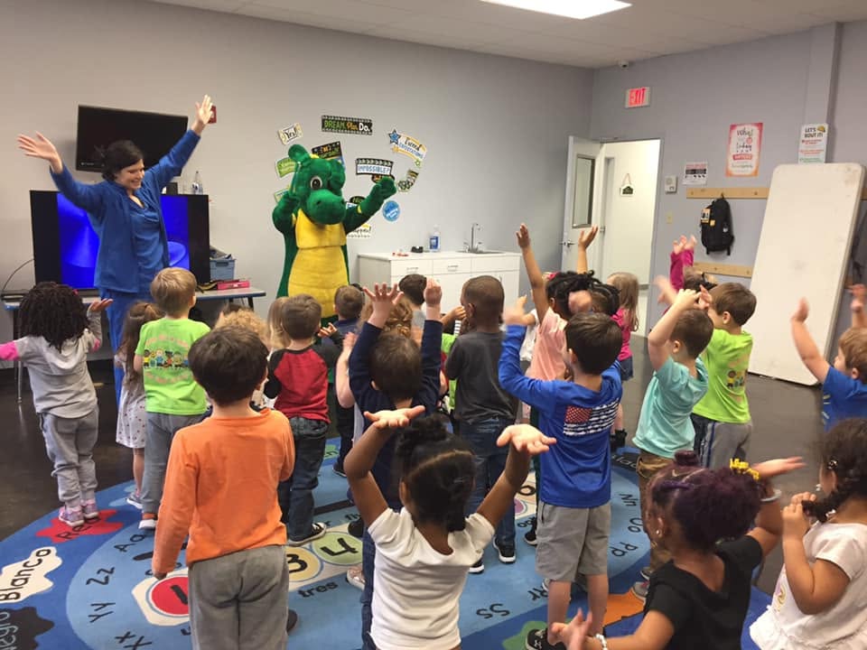Group of children and an adult with a green mascot dancing in a classroom