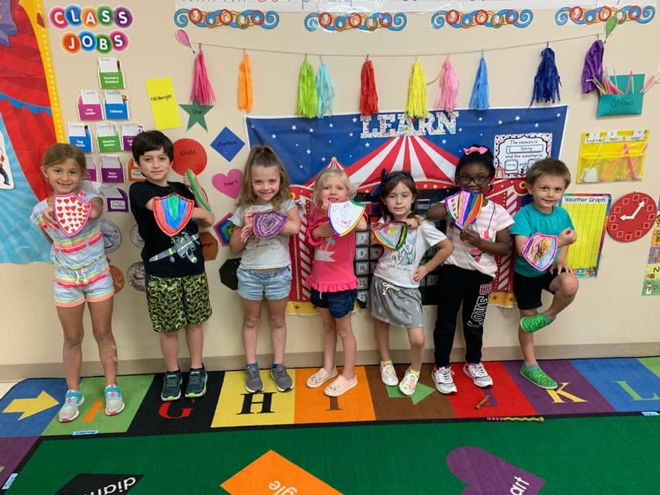 Seven children standing in a classroom holding handmade paper shields