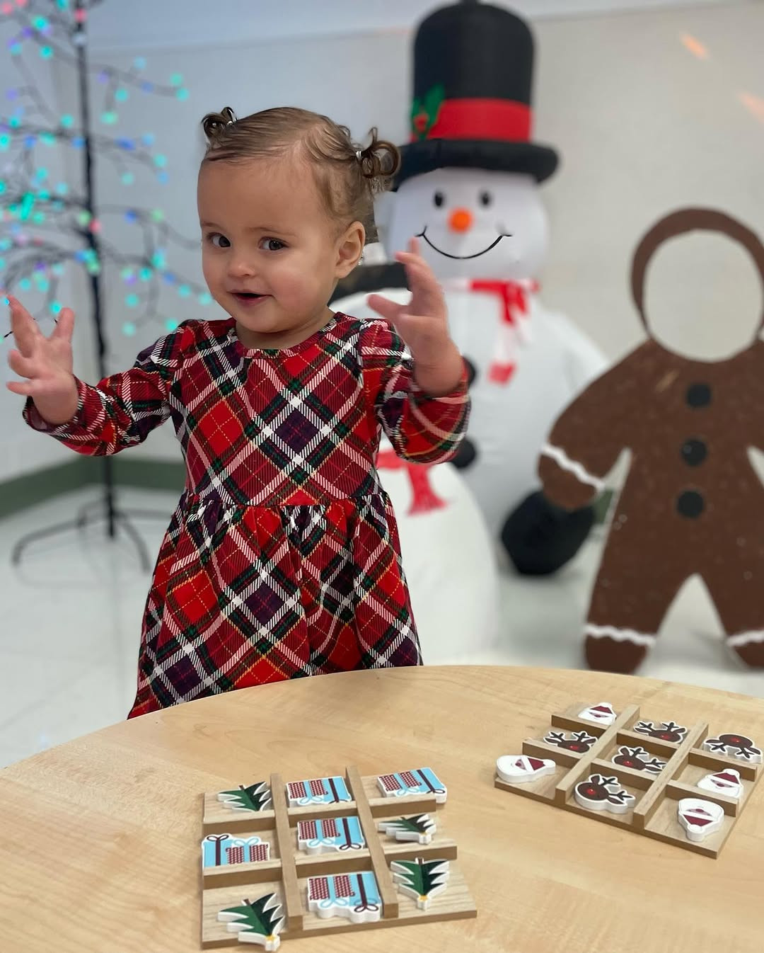 Child in plaid dress with hands raised, standing near a table with games