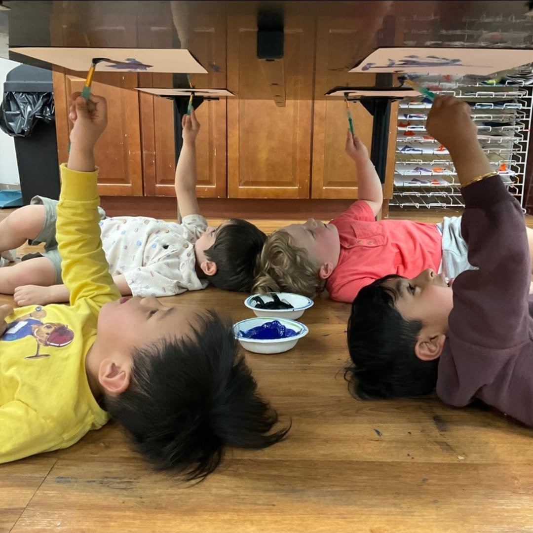 Four children lying on the floor painting on the underside of a table