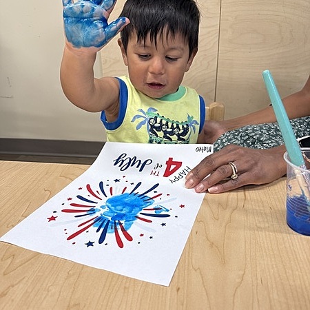 Child with blue paint on hand making a craft, text "Happy 4th of July"