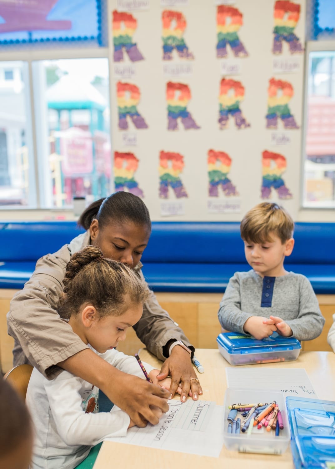 Adult helping a child write at a table with another child nearby in a classroom