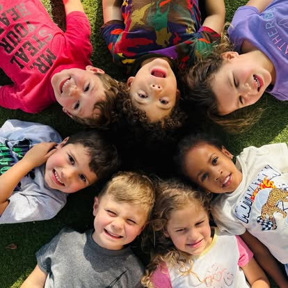 Seven children lying in a circle on grass, looking up