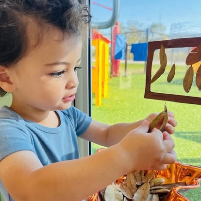 Child holding a leaf next to a window with a playground outside