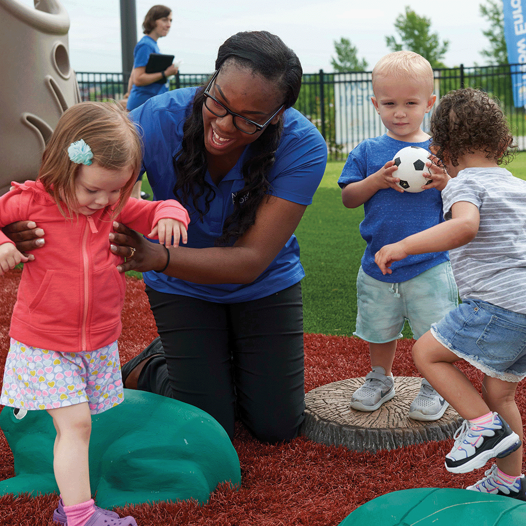 Adult helping a child walk on a playground with two other children nearby