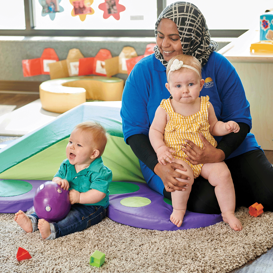 Person sitting on floor holding a child, another child playing with a ball