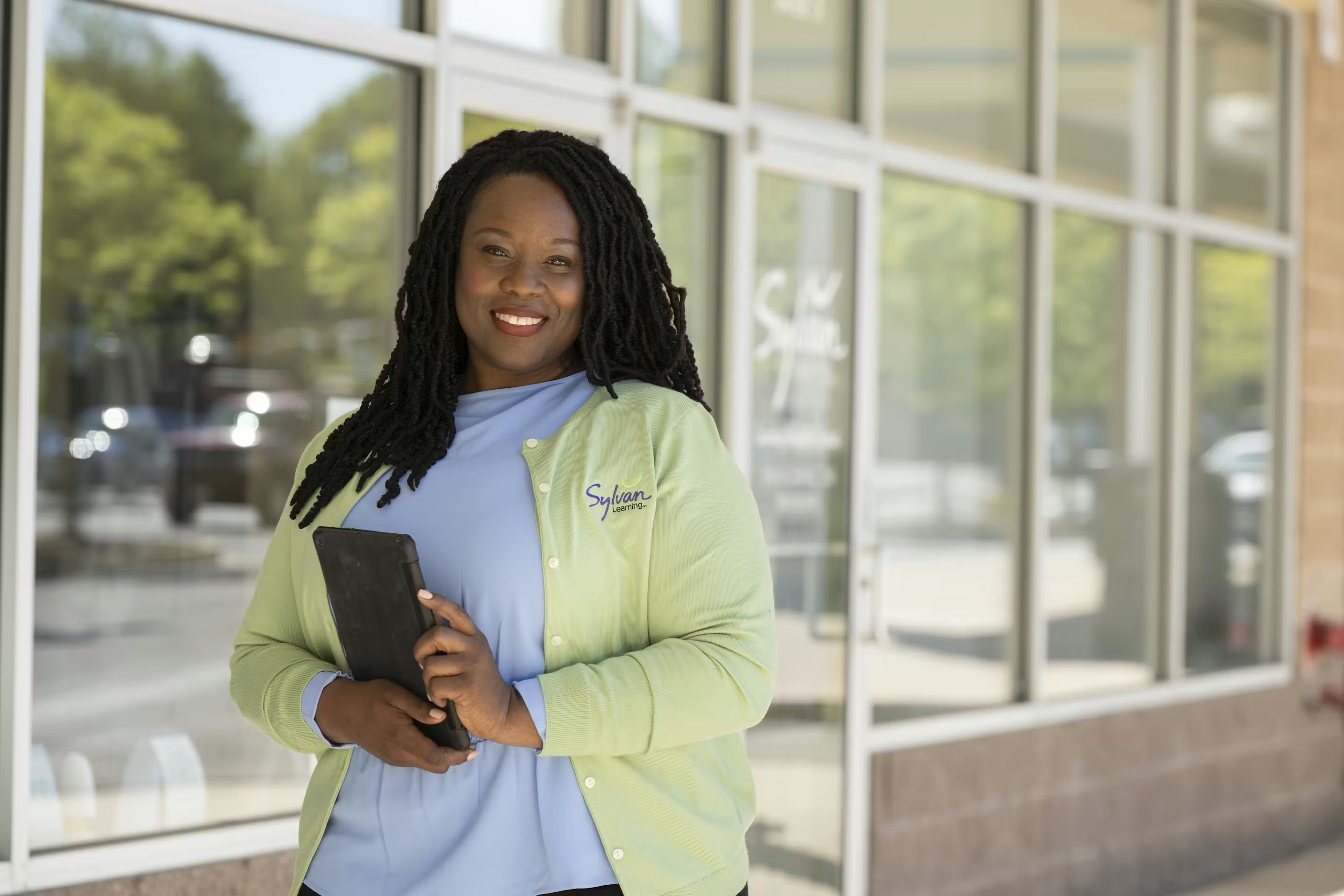 Person with dreadlocks, wearing a light blue top and green cardigan, holding a tablet