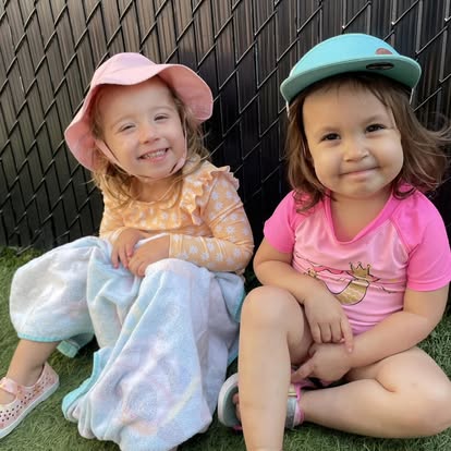Two children wearing hats sitting on grass in front of a fence