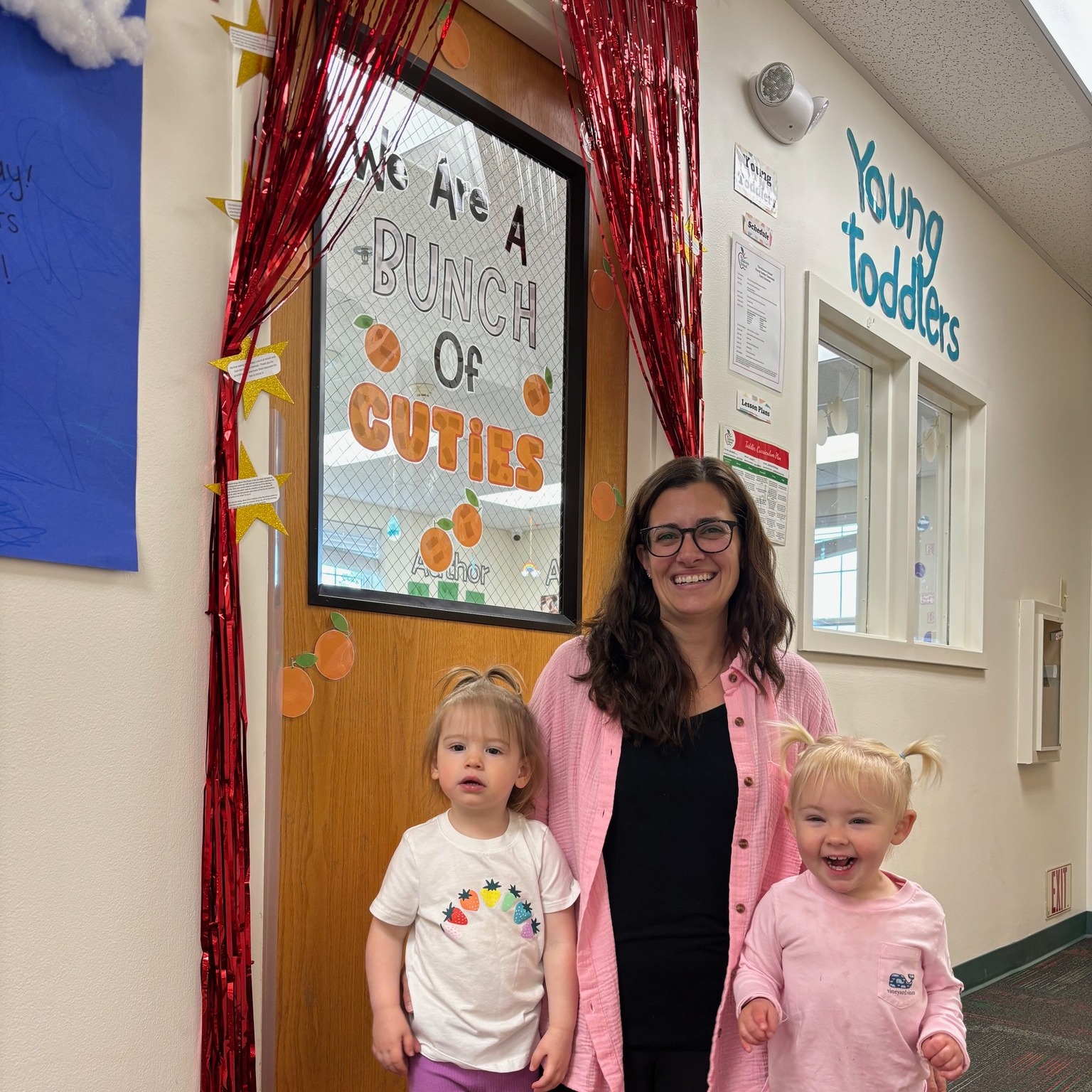 Person with two children in a classroom doorway with a sign that reads "Young Toddlers"