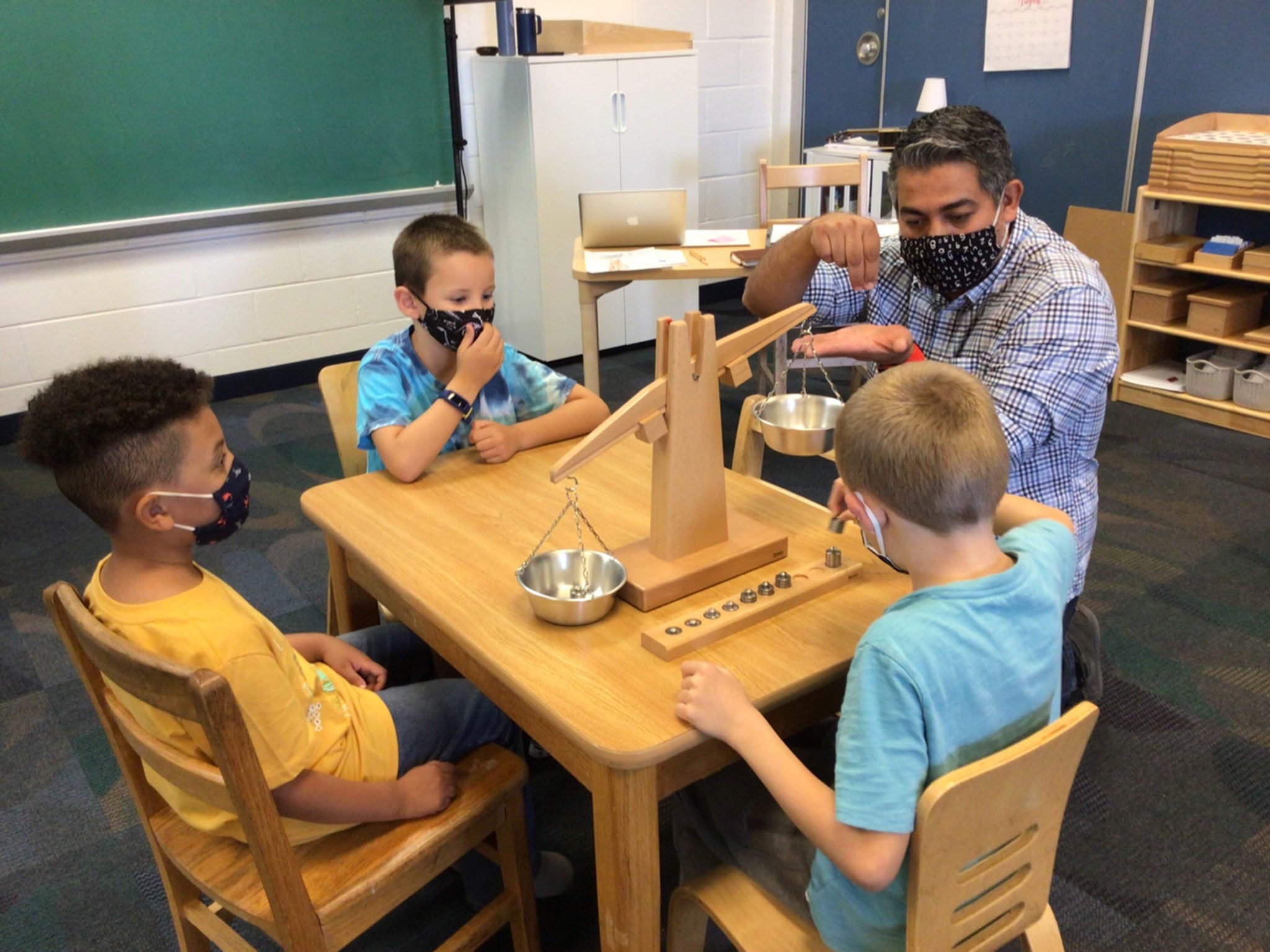 Adult and three children in masks using a wooden balance scale at a table