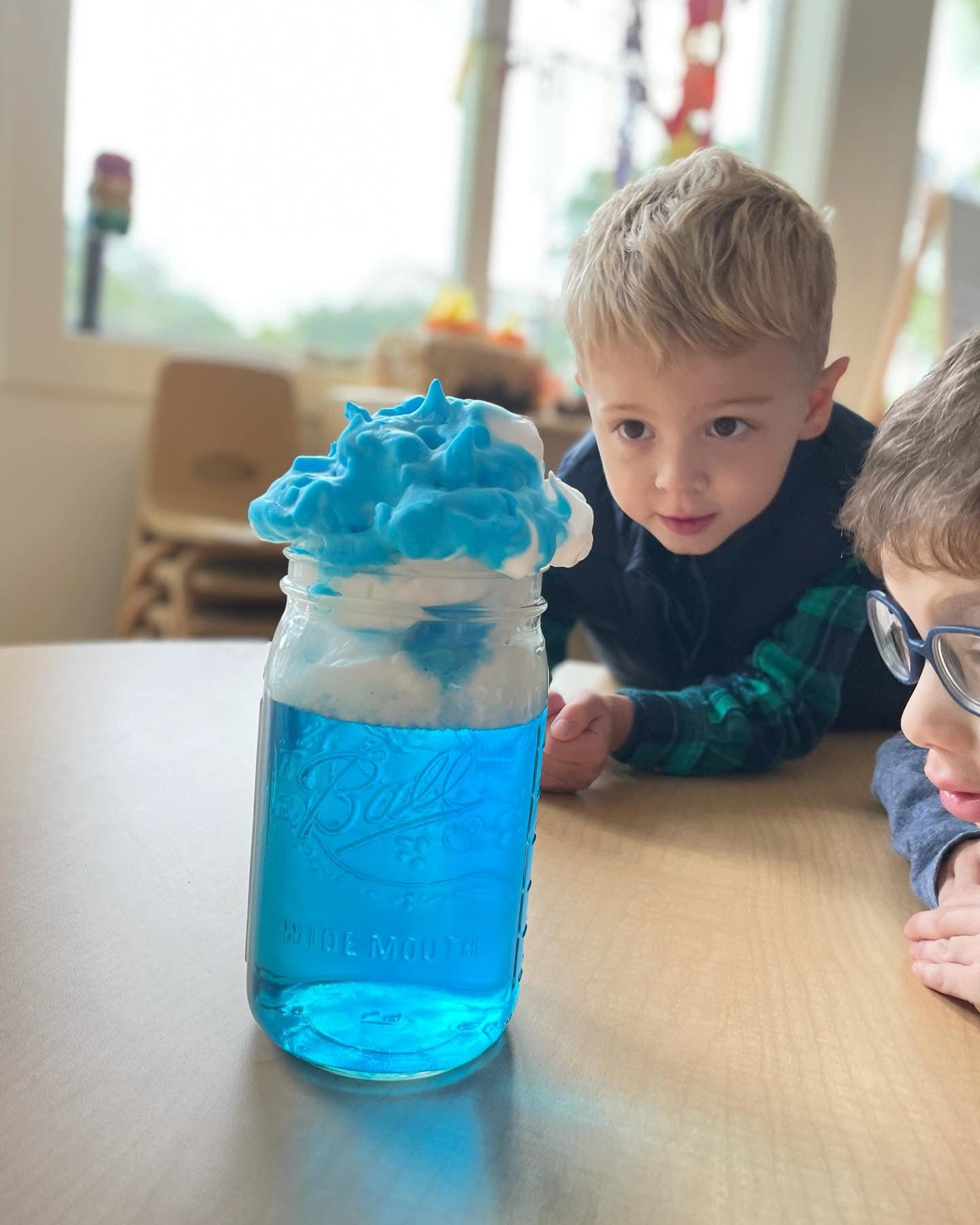 Two children watching a science experiment with blue liquid and foam in a jar