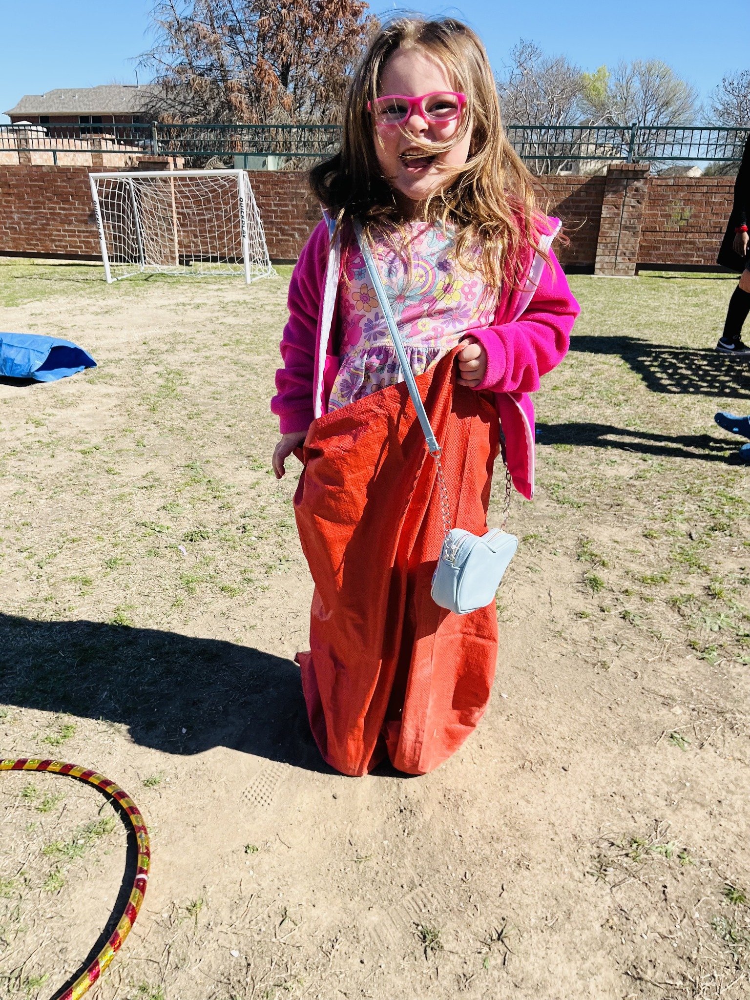 Child in pink glasses standing in a red sack on a grassy field