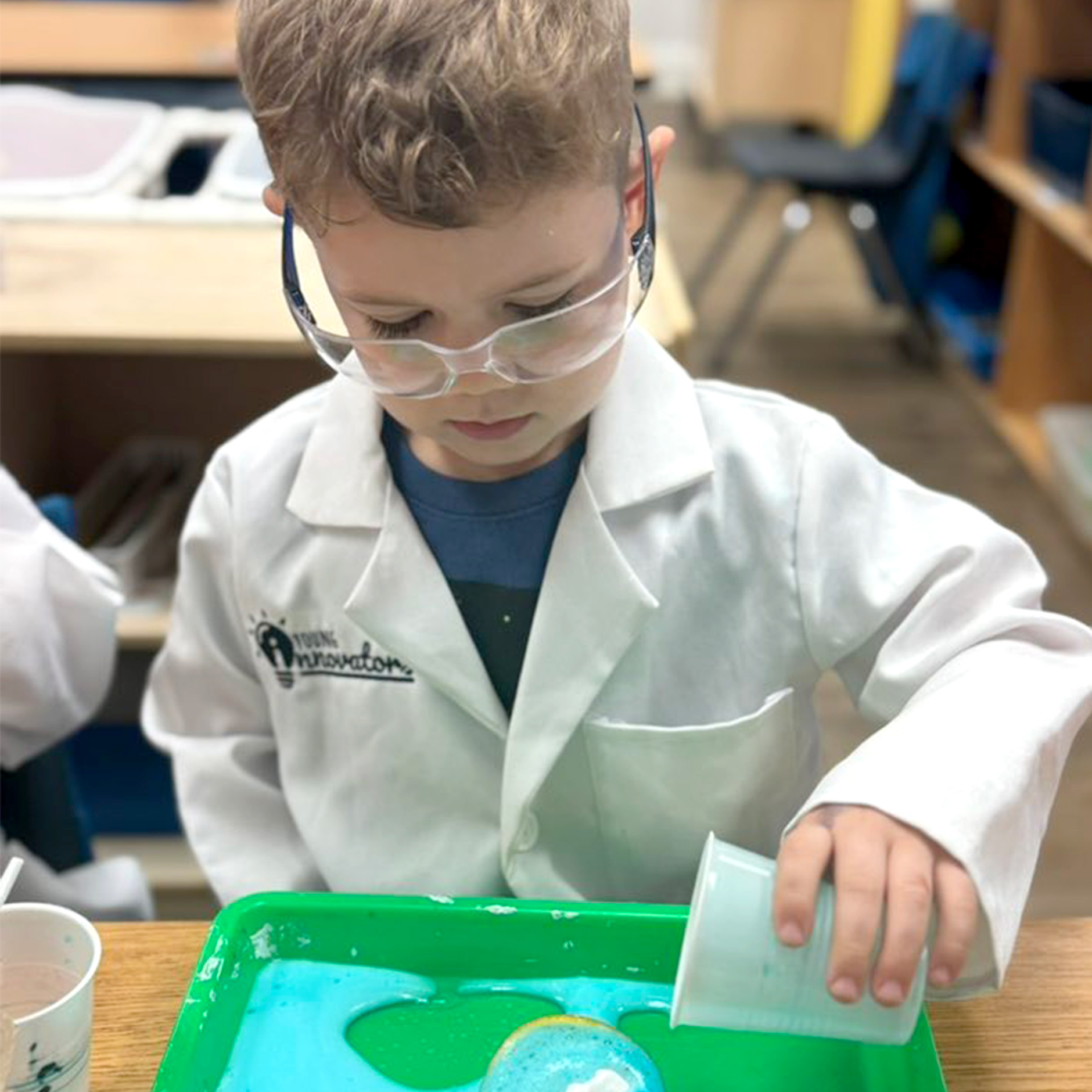 Child wearing a white lab coat and safety goggles pouring liquid into a tray