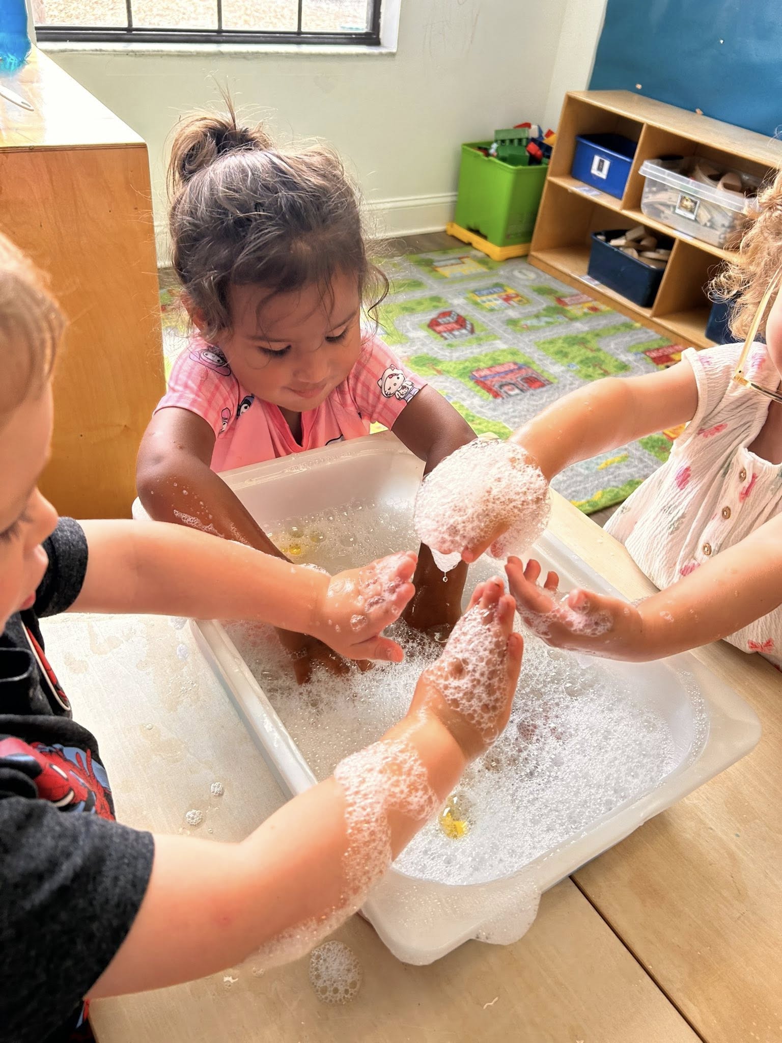 Three children playing with soapy water in a plastic bin at a table