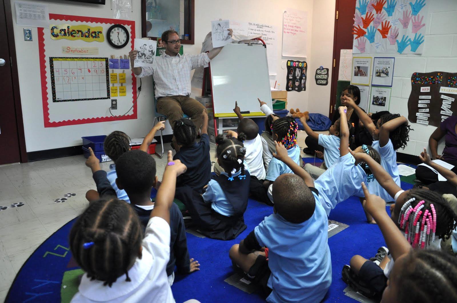 Adult holding papers in front of children sitting on a rug in a classroom