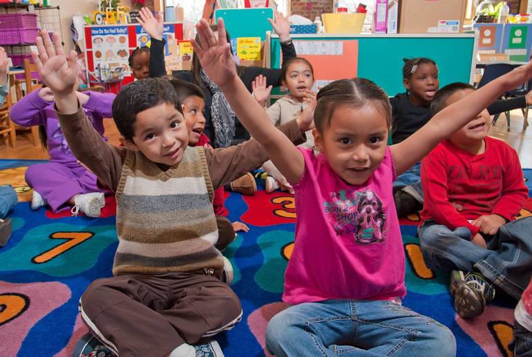 Children sitting on a rug with their hands raised in a classroom