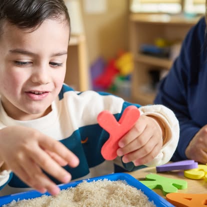 Child holding a red letter shape over a tray of rice