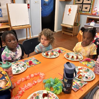 Three children sitting at a table with plates of fruit skewers