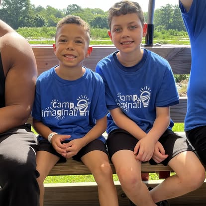 Two children in blue Camp Imagination t-shirts sitting on a bench