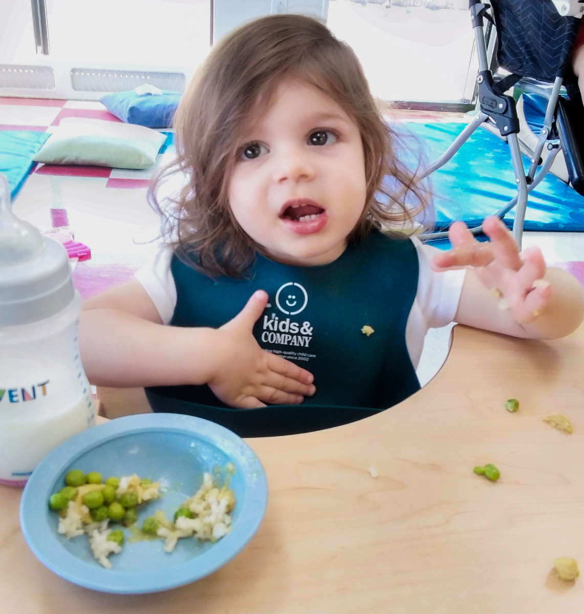 Child in a high chair wearing a bib, with a plate of food and a bottle