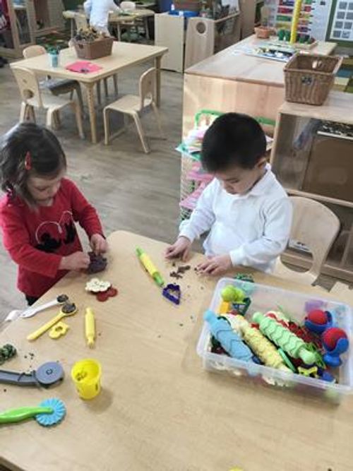 Two children playing with modeling clay and tools at a table in a classroom