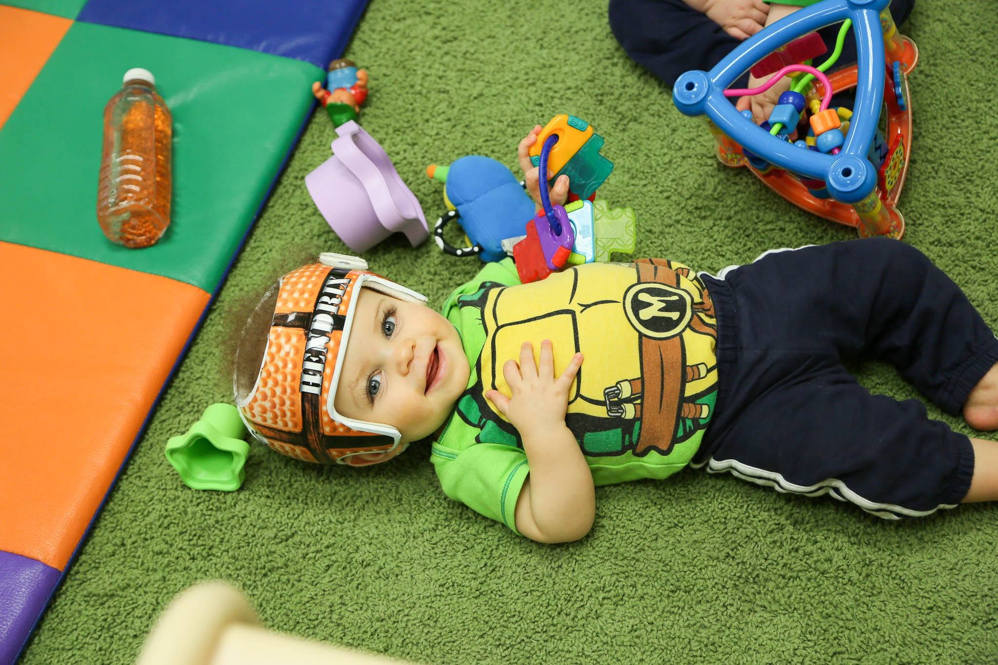 A child lying on a green mat playing with toys while wearing a cranial helmet