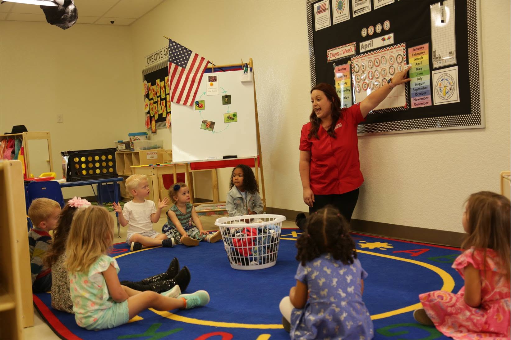 Teacher pointing to a wall calendar while children sit in a circle in classroom