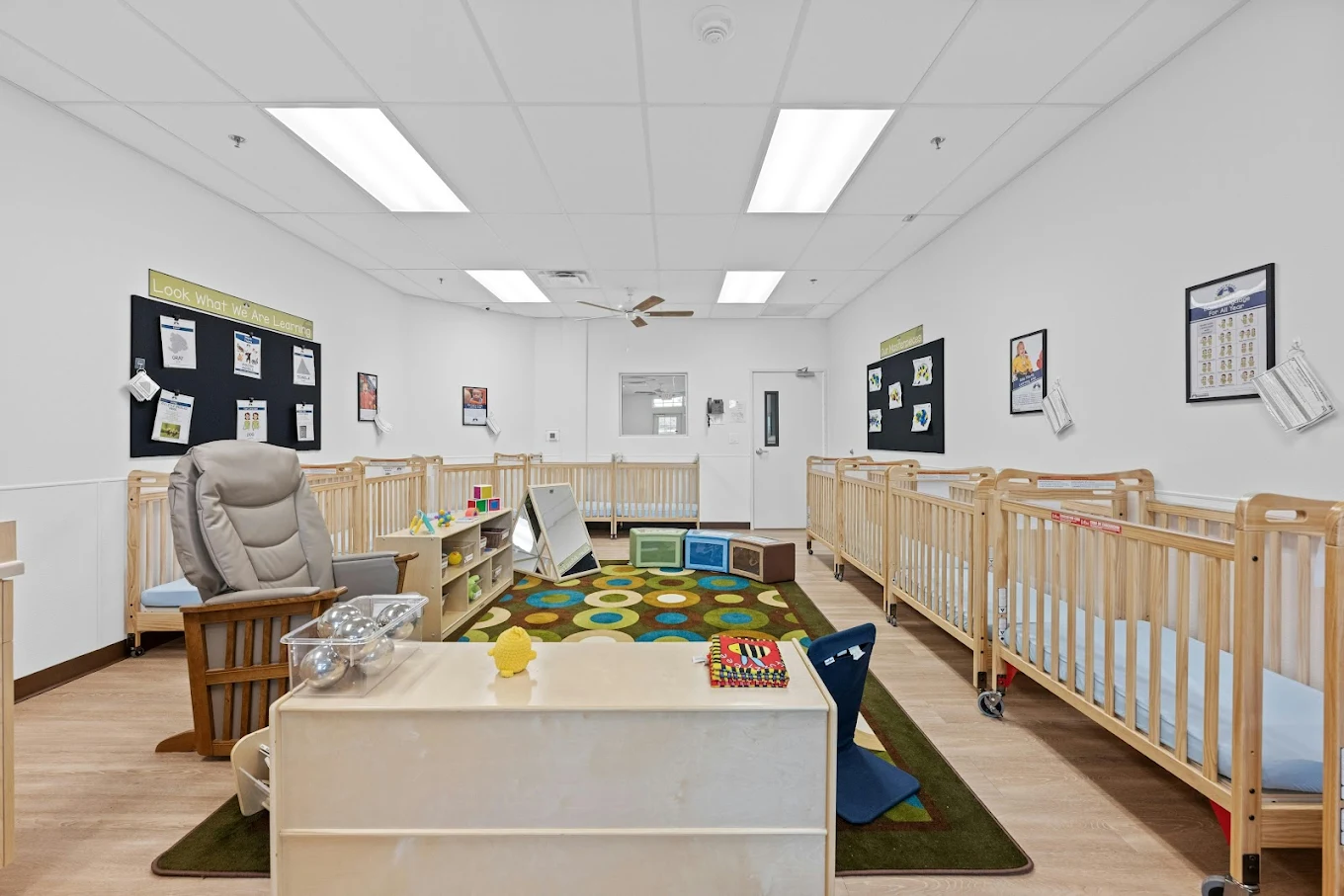Infant classroom with rows of cribs, a play rug, and furniture