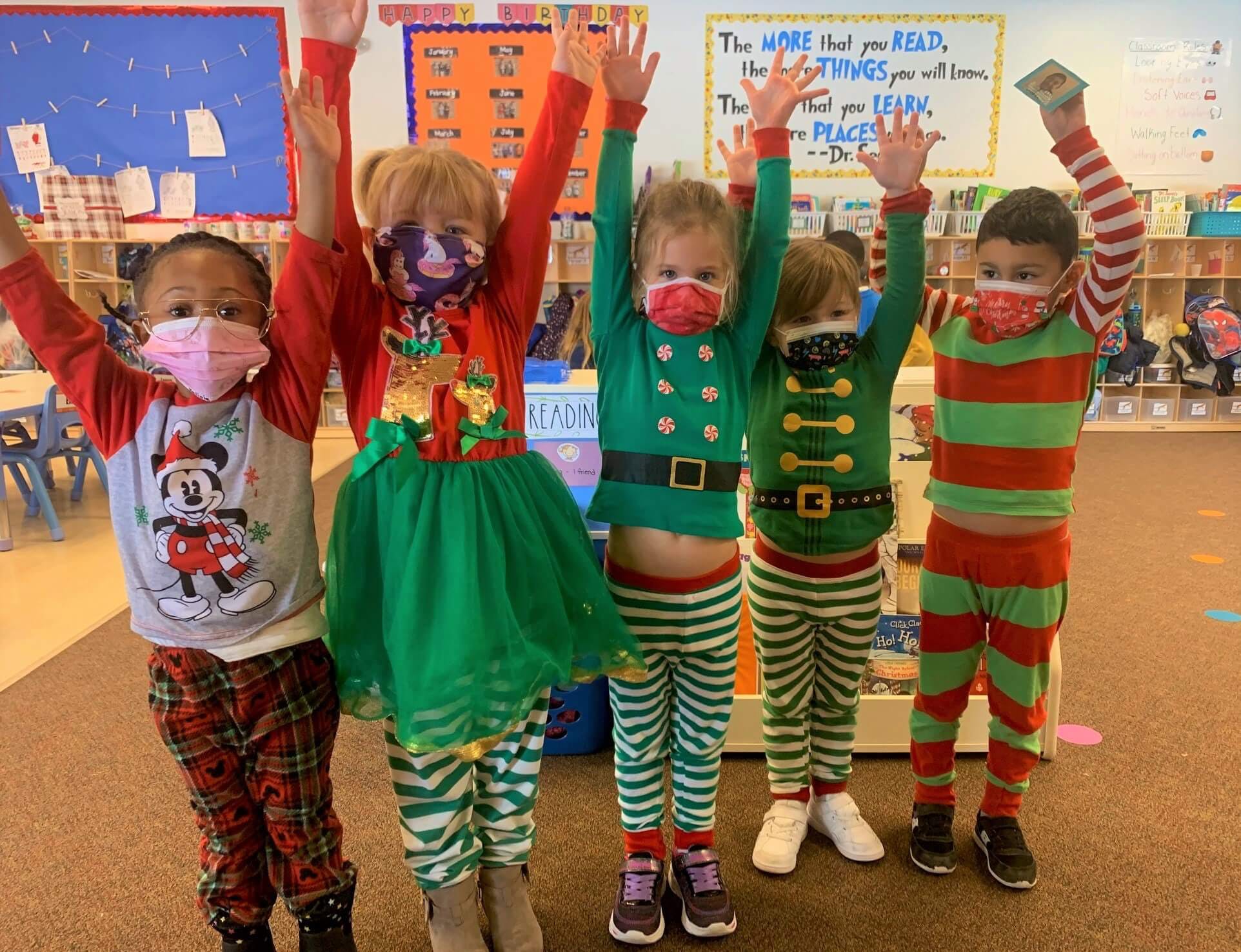 Five children in holiday outfits with arms raised in a classroom