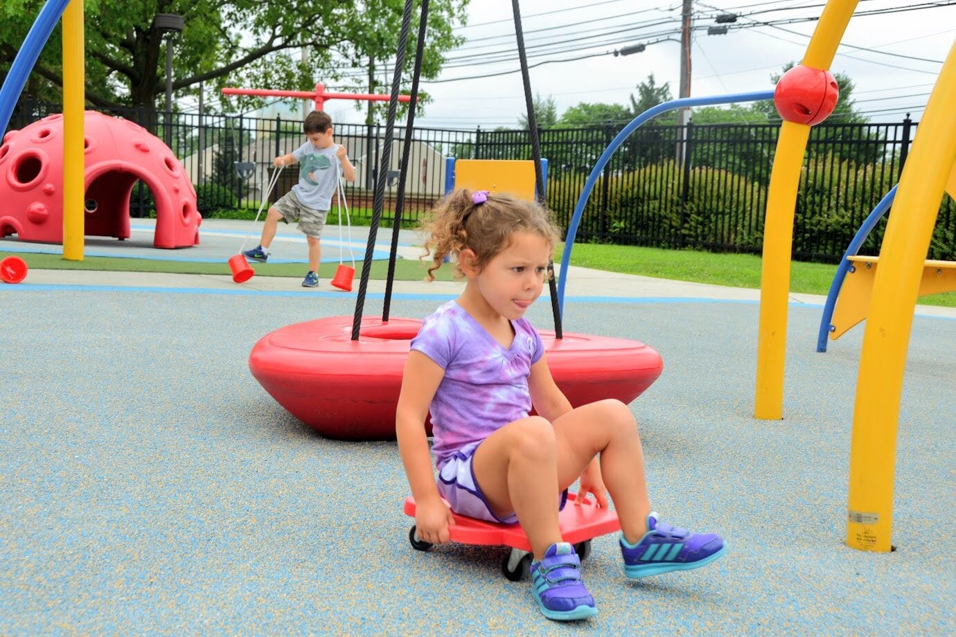 Child sitting on a scooter board on a playground, another child in background