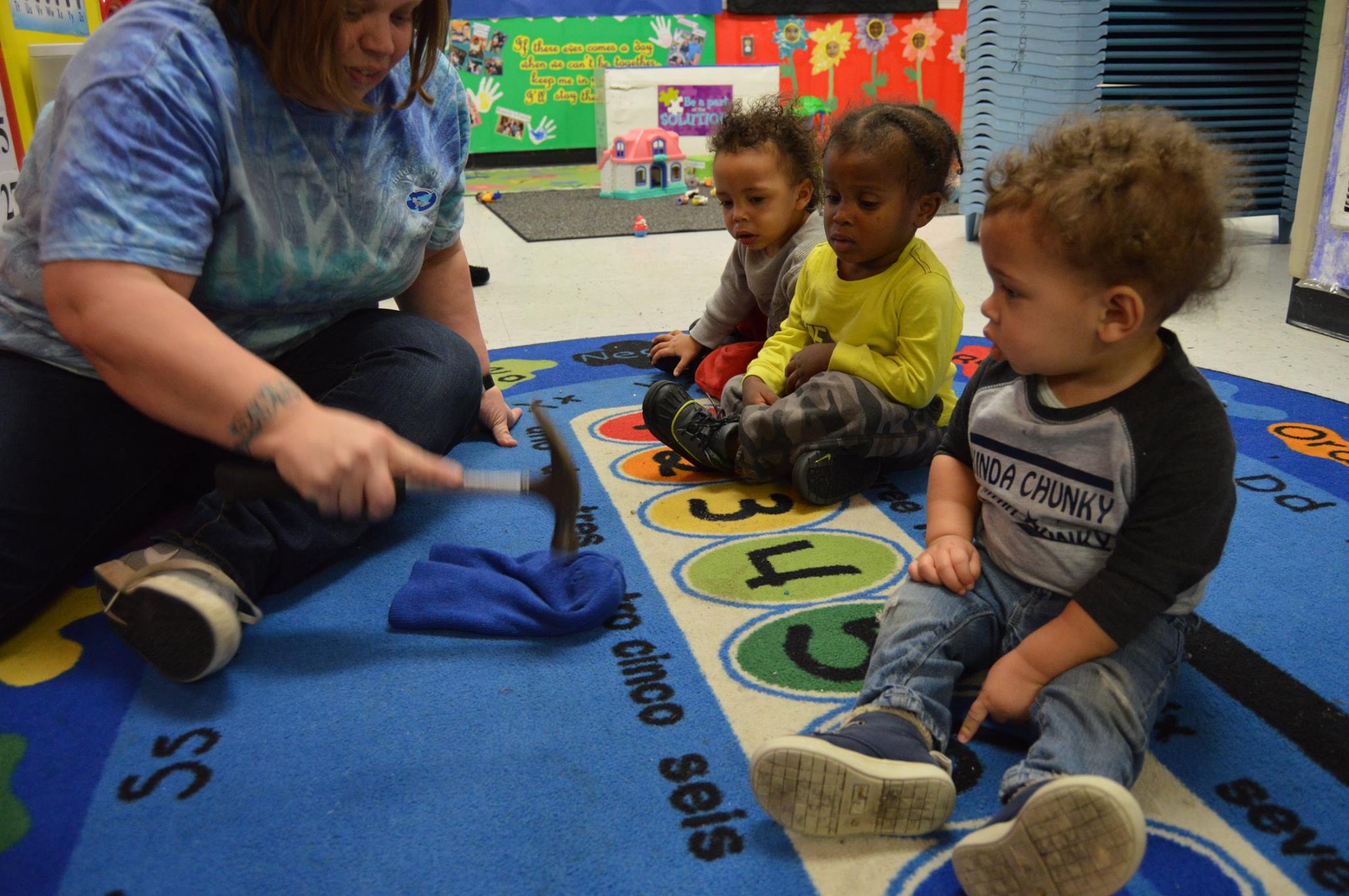 Person showing three children a hammer and blue cloth on a rug