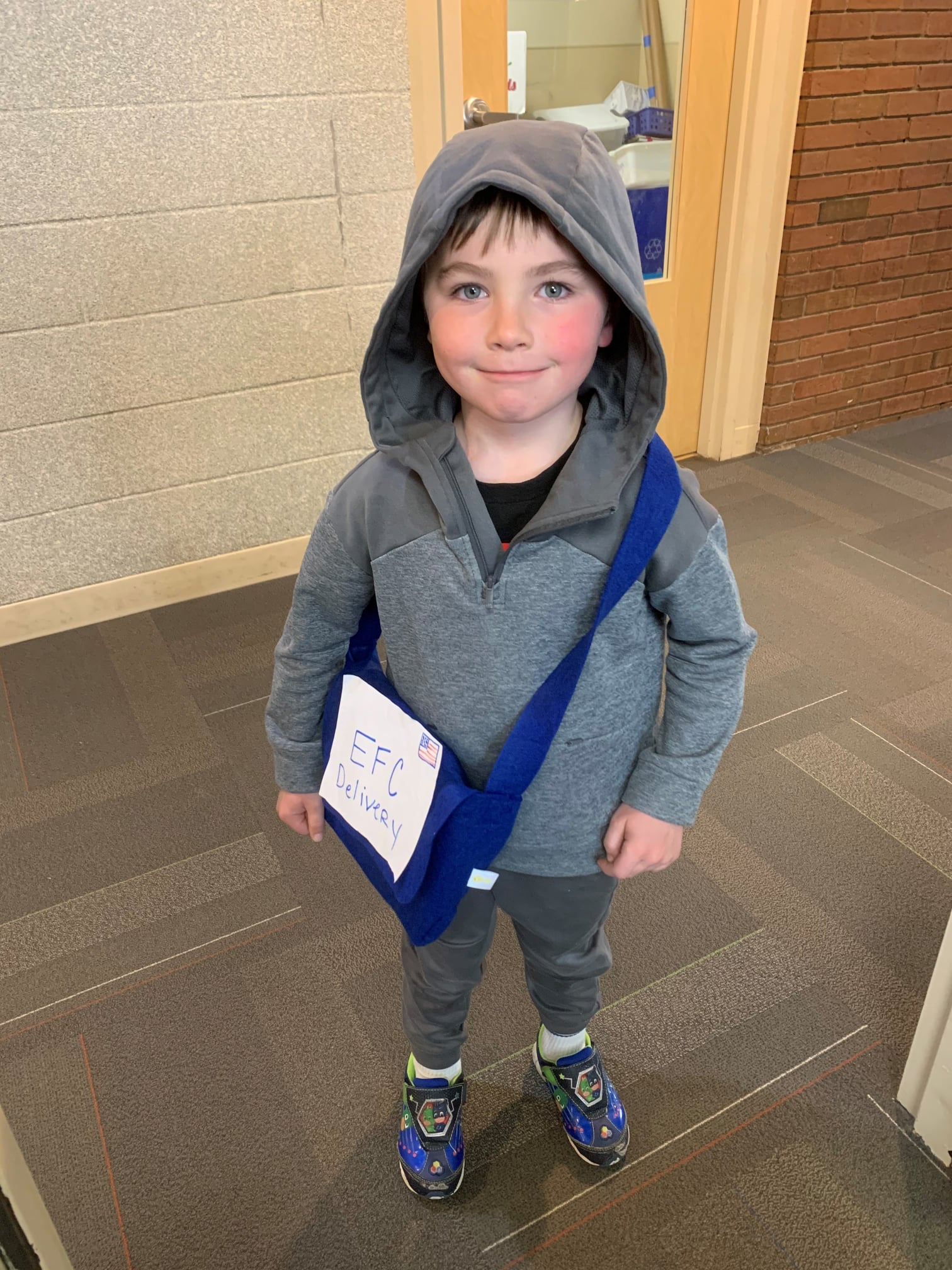 Child wearing a gray hoodie and a blue messenger bag with "EFC Delivery" written on it