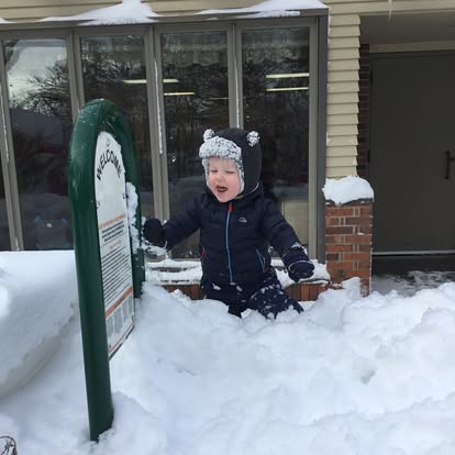 Child in winter clothing playing in deep snow next to a sign