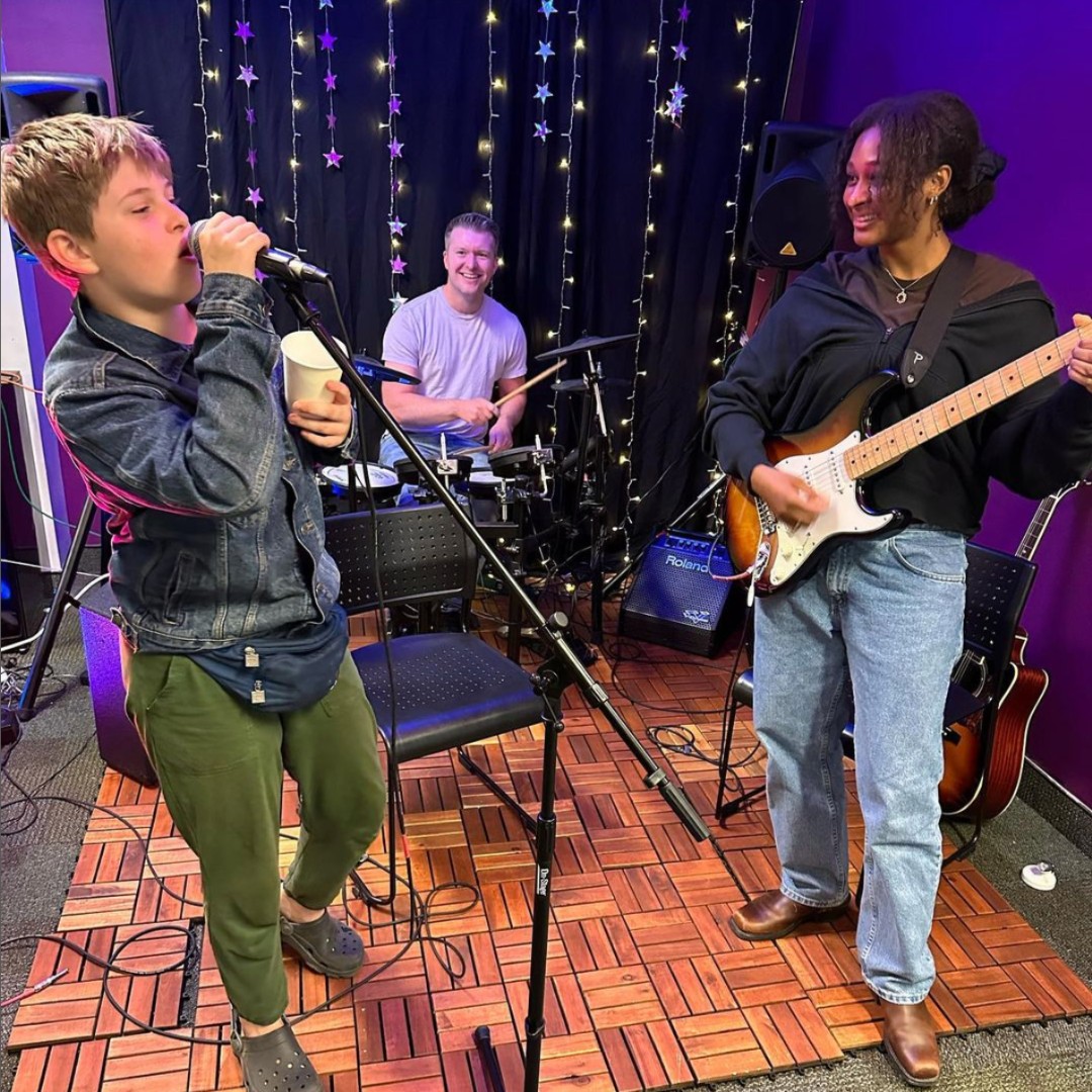Child singing into a microphone, person playing guitar, person playing drums