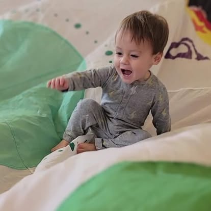 Child in gray pajamas sitting on a bed with mouth open and arm extended