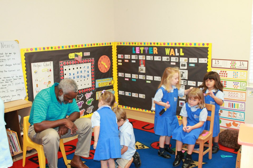 Man sitting on a small yellow chair surrounded by several young children in a classroom