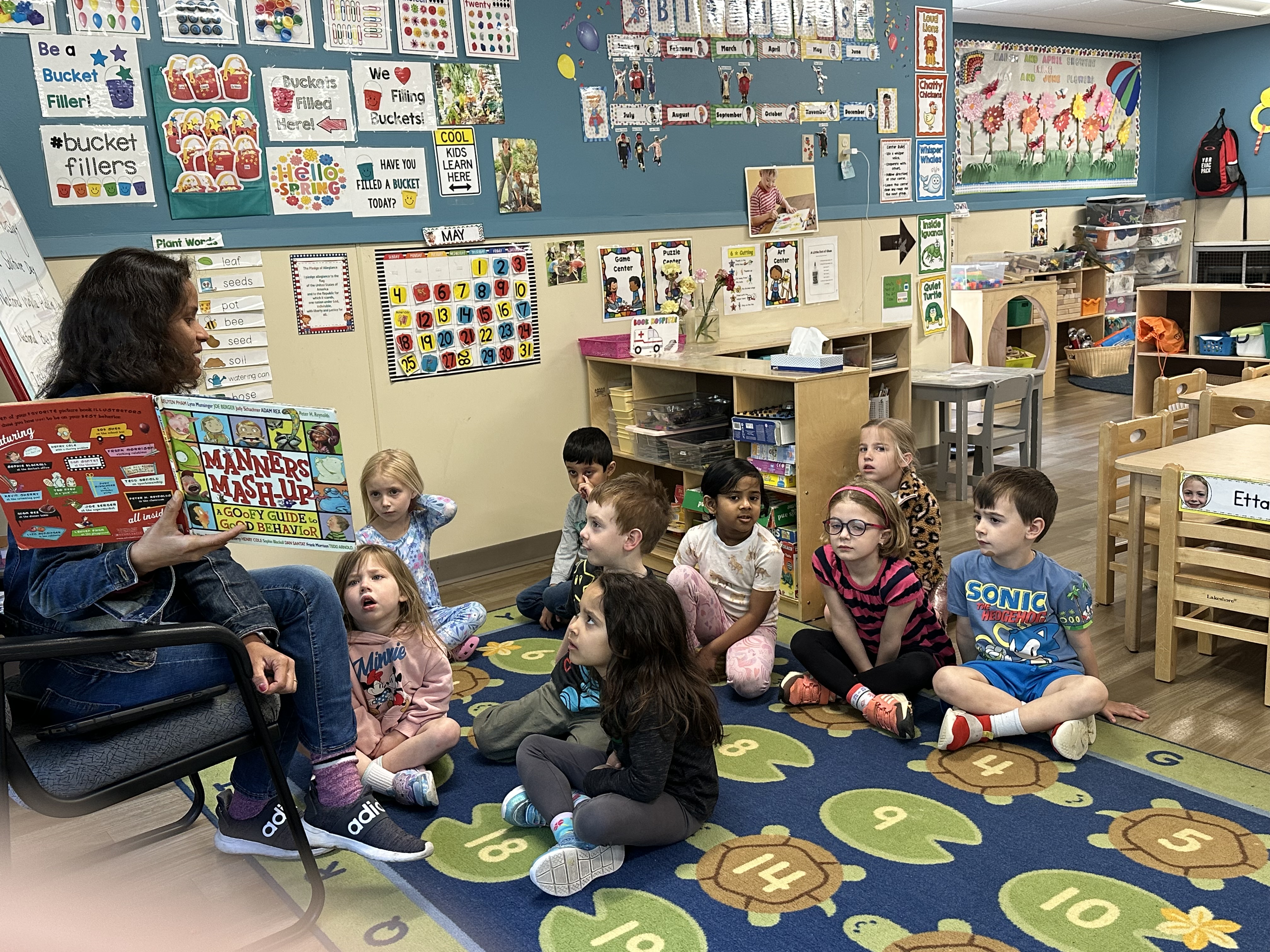 Person reading a book titled "Manners Mash-Up" to children in a classroom