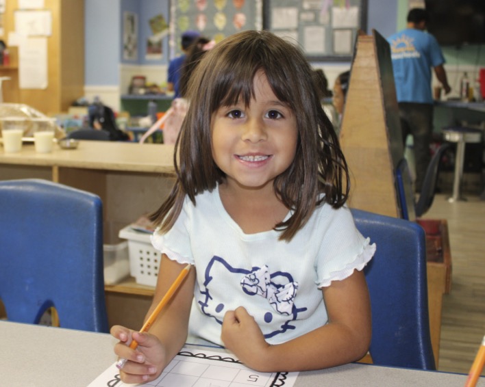 Child smiling while holding a pencil in a classroom